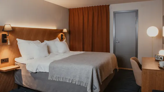 Moderni ja kutsuva hotellihuone, jossa on mukava sänky puisella sängynpäädyllä, lämmin valaistus ja toimiva työpöytä.