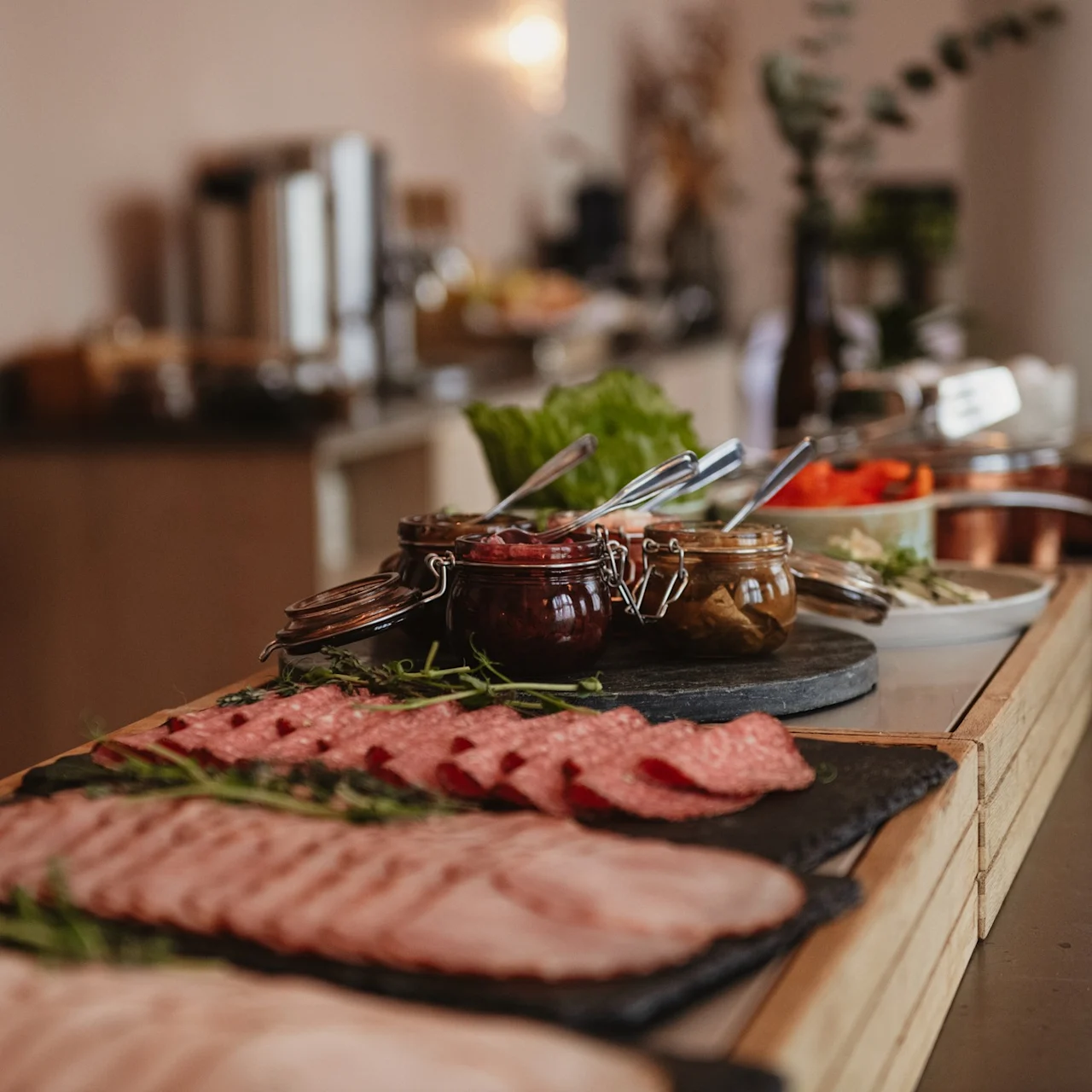 Assorted cold cuts and condiments displayed on wooden platters, garnished with herbs, in a buffet setting with blurred background elements.