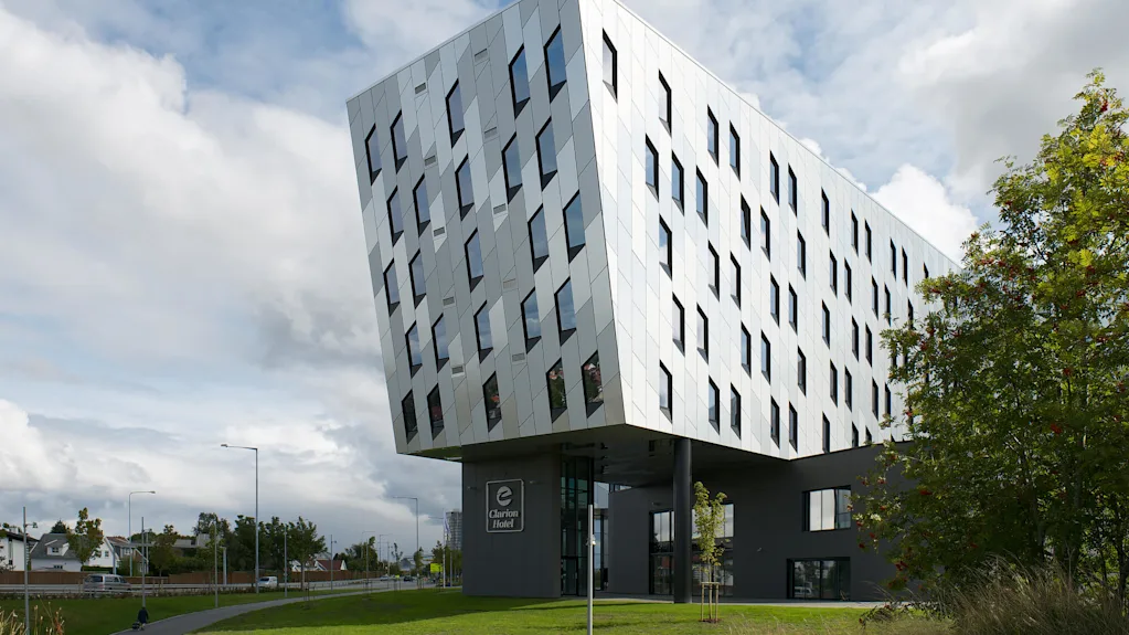 En moderne, vinklet bygning hælder over sin base understøttet af søjler. Facaden har mange vinduer. "Clarion Hotel" er synlig på jordniveau. Omgivet af grønt græs og træer.
