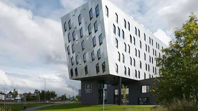 A modern, angular building tilts over its base supported by columns. The facade features numerous windows. "Clarion Hotel" is visible on the ground level. Surrounded by green grass and trees.