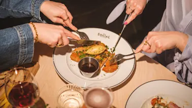 Hands cutting a shared meal on a plate with the word SOCIAL, enjoying a dining experience together.