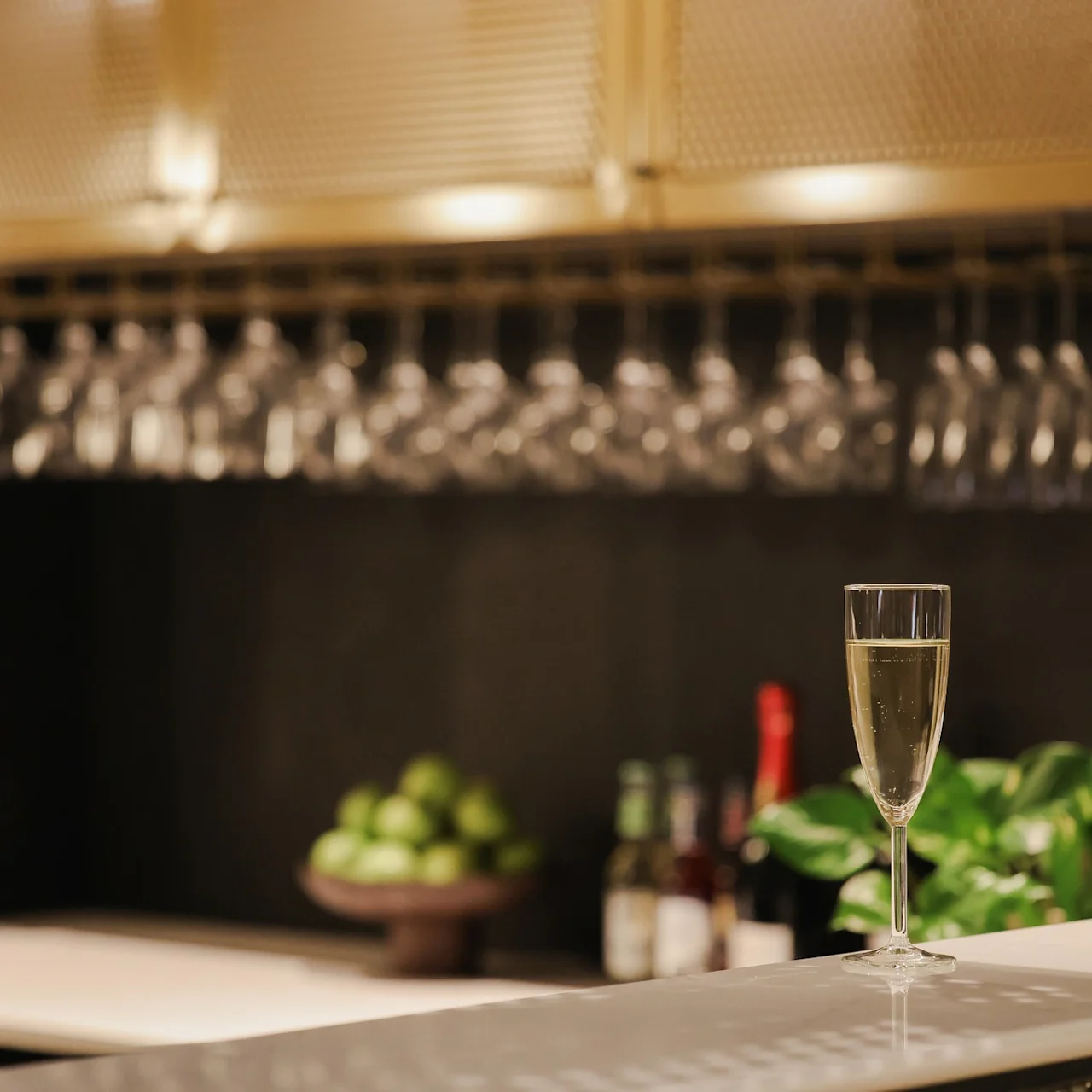 A glass of champagne sits on a bar counter, surrounded by a bowl of green apples, bottles, hanging glassware, and vibrant green plants.