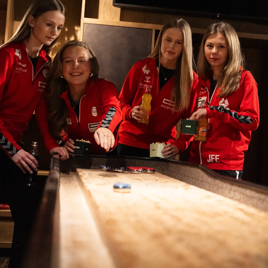 People in red shirts playing shuffleboard, holding drinks and popcorn. Text on popcorn boxes says "make", and "JFE" on a shirt.