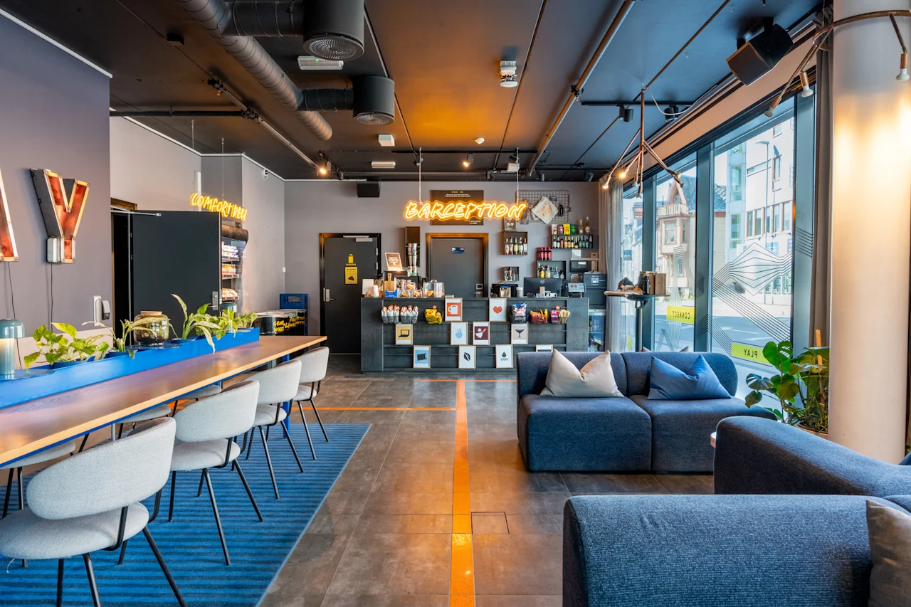 En moderne hotellobby med stilfulde møbler og indretning. Reception har neonskiltet "CAFEPTION", omgivet af udsigt til byen gennem store vinduer, polstrede siddepladser og moderne pendelbelysning.