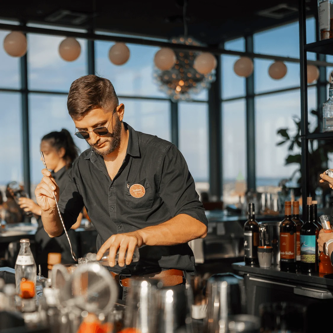 En bartender rører en drink i en tagbar, mens sollys strømmer gennem store vinduer. Flasker, glas og frugter er arrangeret på disken; kunder taler i baggrunden.