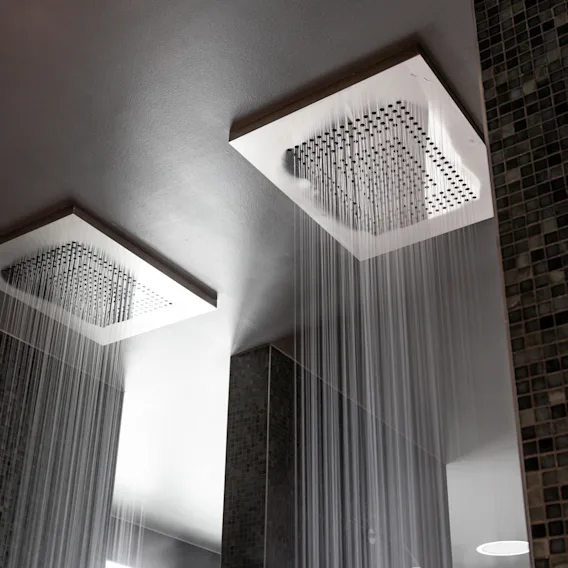 Two ceiling-mounted square showerheads releasing water streams in a modern bathroom with dark tiled walls.