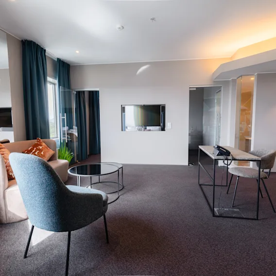 Et moderne rom med en grå lenestol og en sofa vendt mot en veggmontert TV, komplettert av et lite rundt bord. Store vinduer med blå gardiner lyser opp rommet.