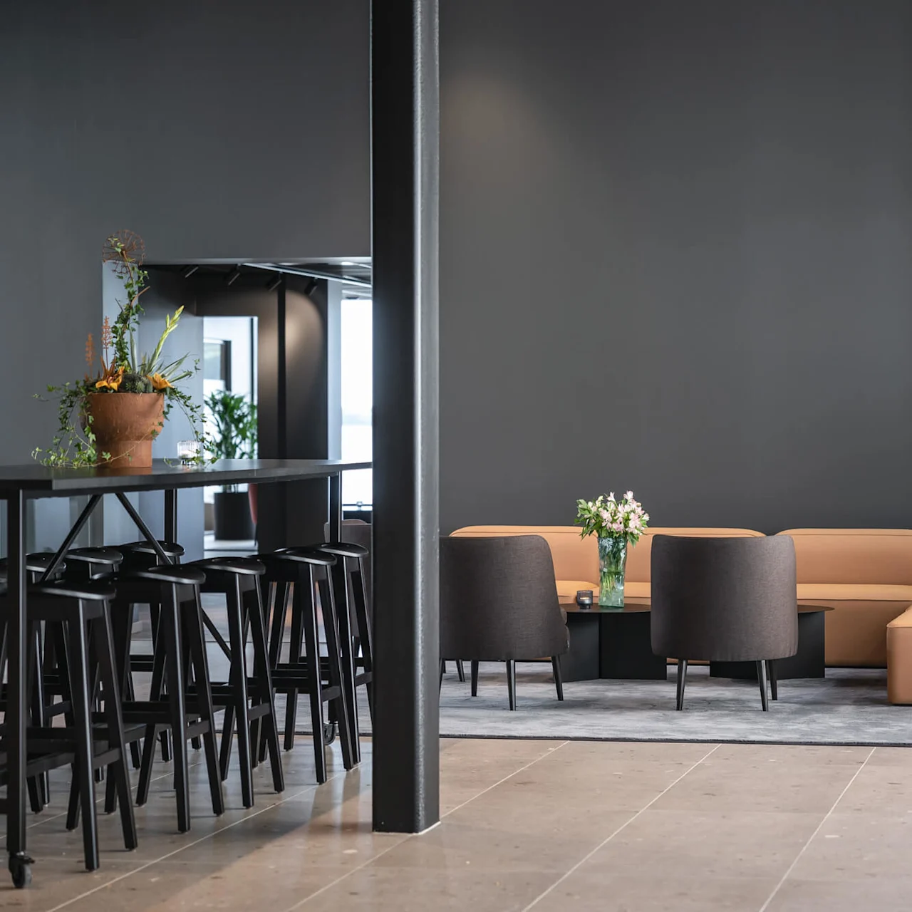 En brun sektionssofa og mørke stole omkranser et bord med blomster i et moderne, minimalistisk rum, med et højt barbord med skamler og potteplanter.