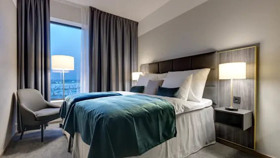 En pent redd seng med grønt teppe står i et moderne hotellrom. Myk belysning fra lamper og et stort vindu skaper en rolig kveldsstemning.