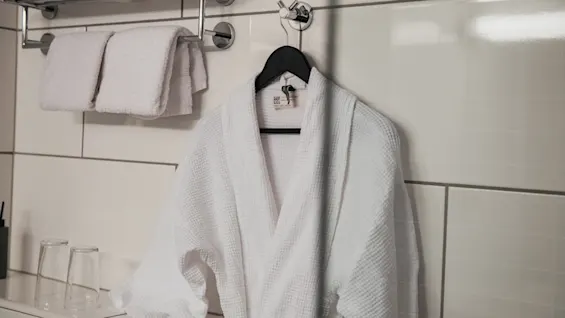En hvid badekåbe hænger på en krog mod en flisevæg, ledsaget af foldede håndklæder på en hylde ovenover og to glas på en bordplade.