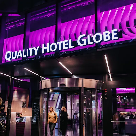 En hotellentré har en roterande dörr med två personer som går ut. Ovanför finns en ljus skylt med texten "QUALITY HOTEL GLOBE" upplyst i vitt mot en livfullt lila fasad.
