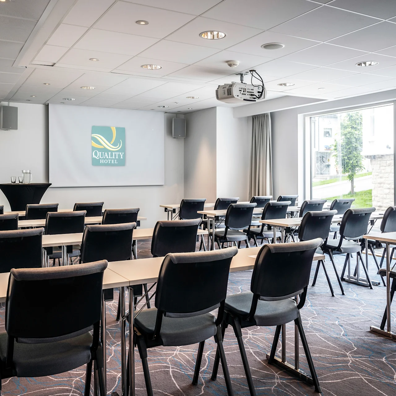 Rader av stolar vetter mot en projektionsskärm med texten "Quality Hotel" i ett väl upplyst konferensrum, med glasfönster som visar en utomhusvy.