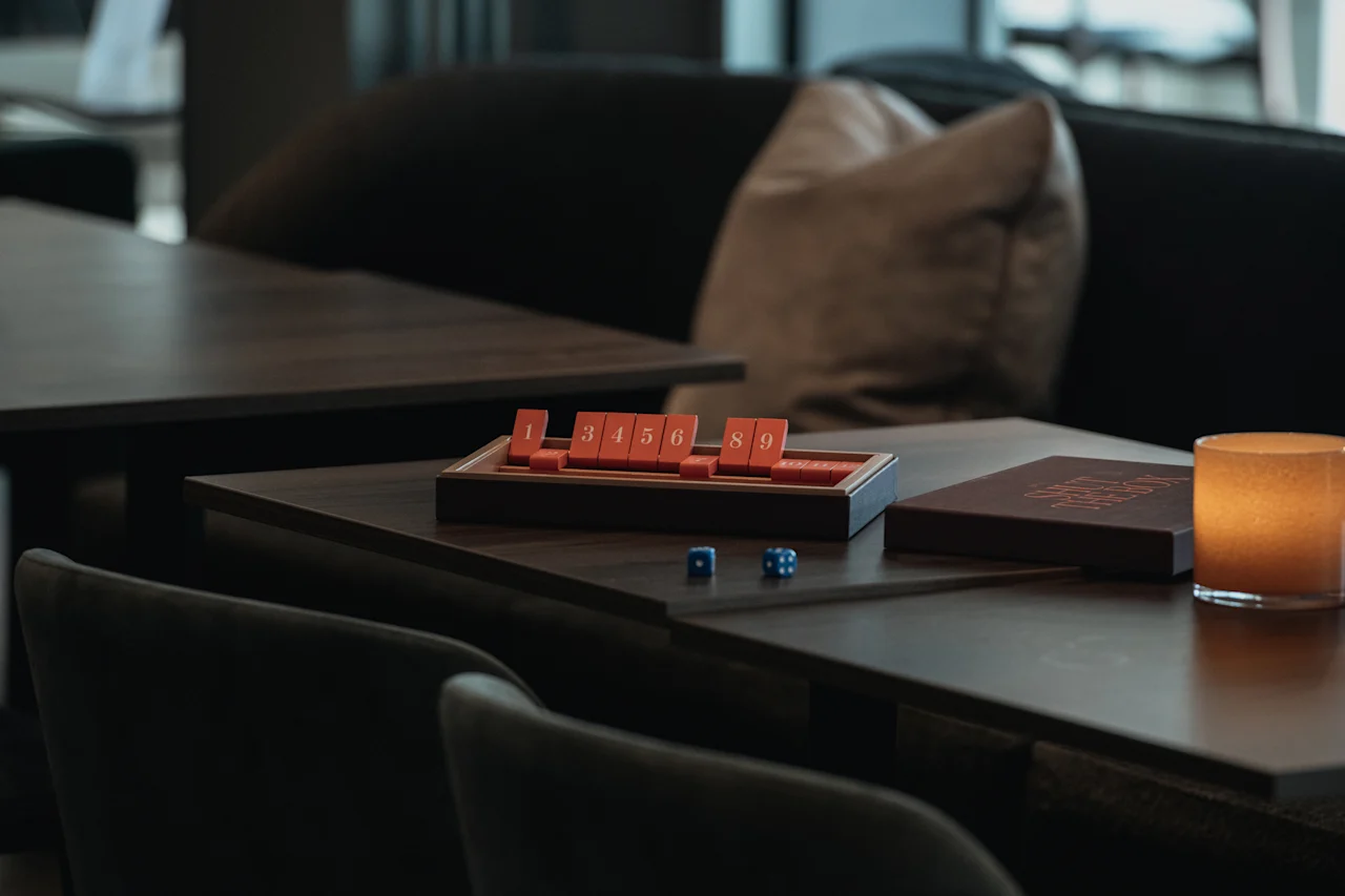 Ett brädspel med numrerade brickor 1 3 4 5 6 8 9 och tärningar på ett bord, bredvid ett ljus. Spellådan säger More Than Just A Game.