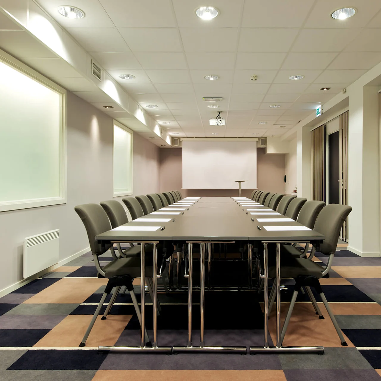 Et langt konferansebord med stoler arrangert symmetrisk; papirer og penner ligger på bordet. Rommet har et projektorlerret og et teppe med firkantede mønstre.