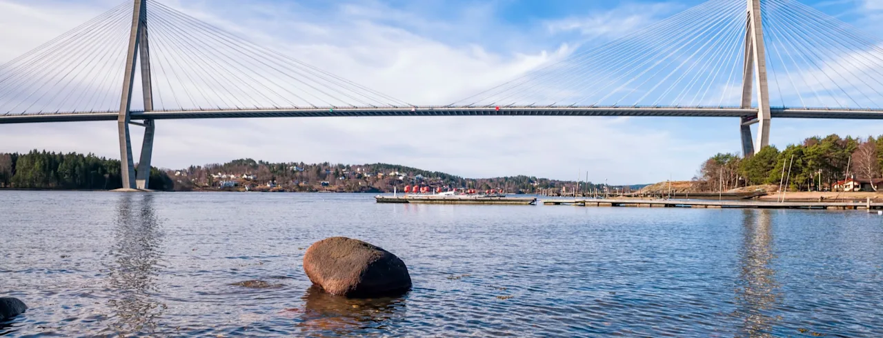 Suspension bridge spans across a wide river, supported by tall towers with cables. The river reflects the bright blue sky, surrounded by distant hills and scattered trees along the banks.