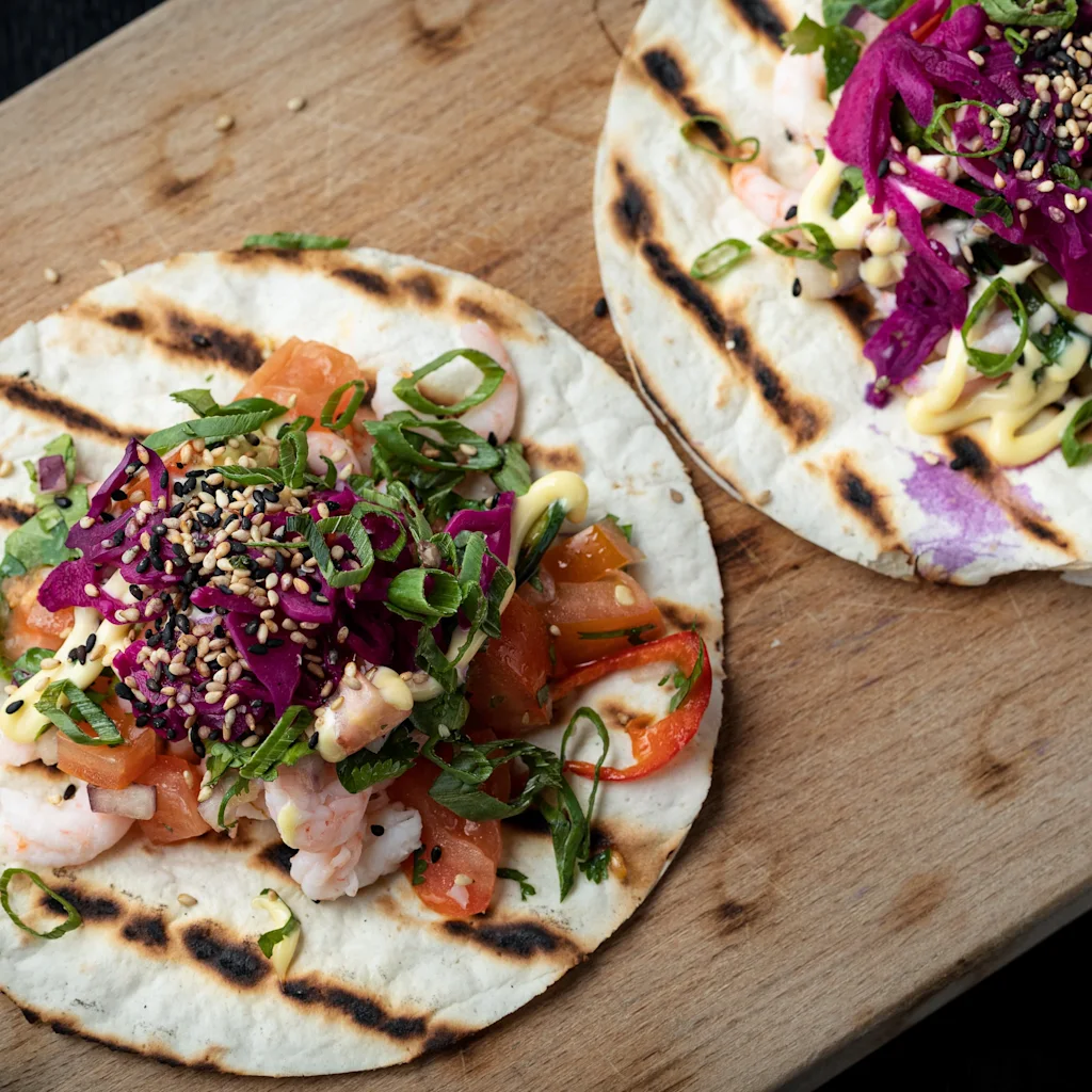 Två grillade tortillor toppade med räkor, tomater, strimlad rödkål, salladslök, sesamfrön och sås på en träbricka.