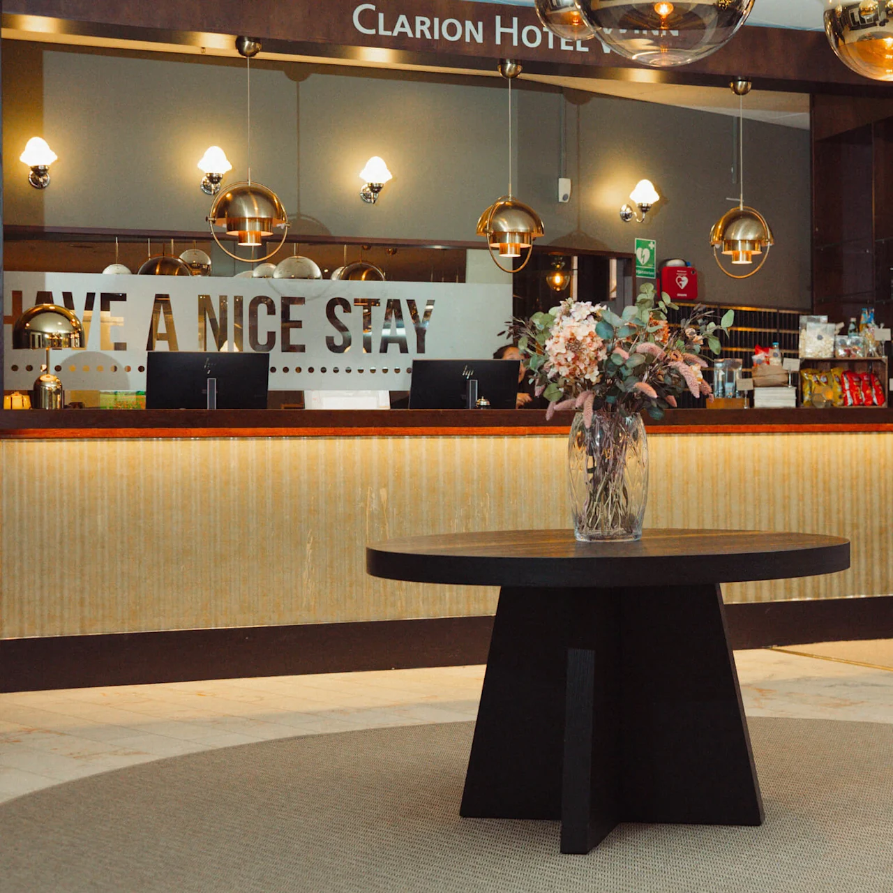 Et rundt bord med en blomsterarrangement står i en hotellobby. Receptionen i baggrunden har computere, snacks og drikkevarer. Tekst: "CLARION HOTEL" og "HAVE A NICE STAY."