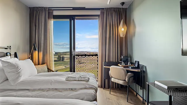 To pent re oppredde enkeltsenger i et hotellrom vender mot et vindu med utsikt over åpne jorder og blå himmel. Ved siden av er et skrivebord med stol og lampe.