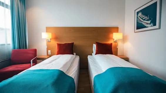 To prydelig redde enkeltsenger med turkise tepper er i et ryddig hotellrom med tregavler, røde puter og nattbordslamper. Et vindu med blå gardiner og et innrammet båtmaleri er til stede.