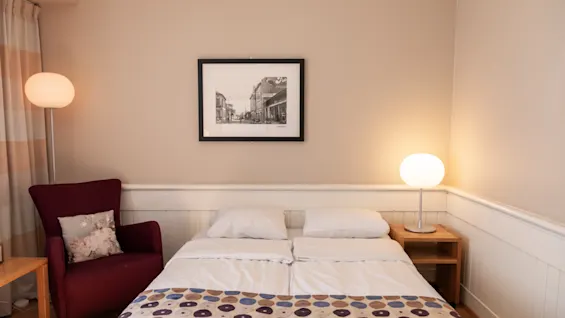 Seng pænt redt med hvidt sengetøj og mønstret plaids; omgivet af natbord med tændt lampe, indrammet sort-hvidt foto på væggen og rød lænestol i hyggeligt, blødt oplyst rum.