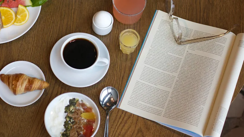 A breakfast spread on a wooden table includes coffee, fruit, yogurt, a croissant, juice, and an open magazine with glasses beside it. A vase with a pink flower adds decoration.
