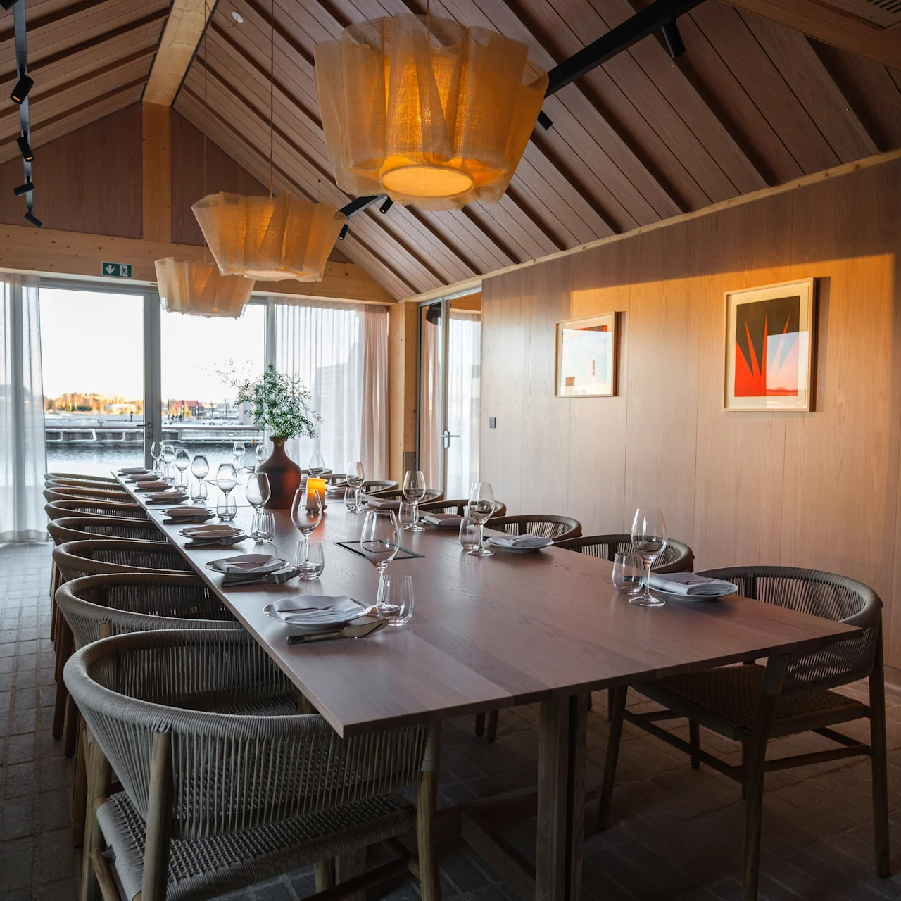 A long wooden dining table is set elegantly with glassware and plates, surrounded by wicker chairs. Soft pendant lights hang above in a rustic room with wooden walls and framed art.