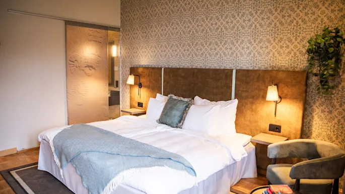 A large bed is covered with white linens and a blue blanket, situated against a patterned wallpaper in a warmly lit bedroom with side tables and a cushioned chair.