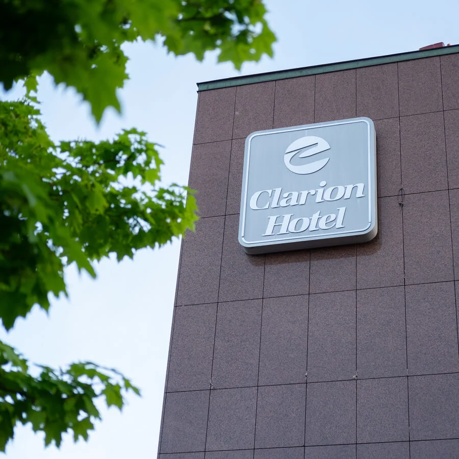 A sign with "Clarion Hotel" is mounted on a brown building facade. Green tree leaves frame the view under a clear blue sky.