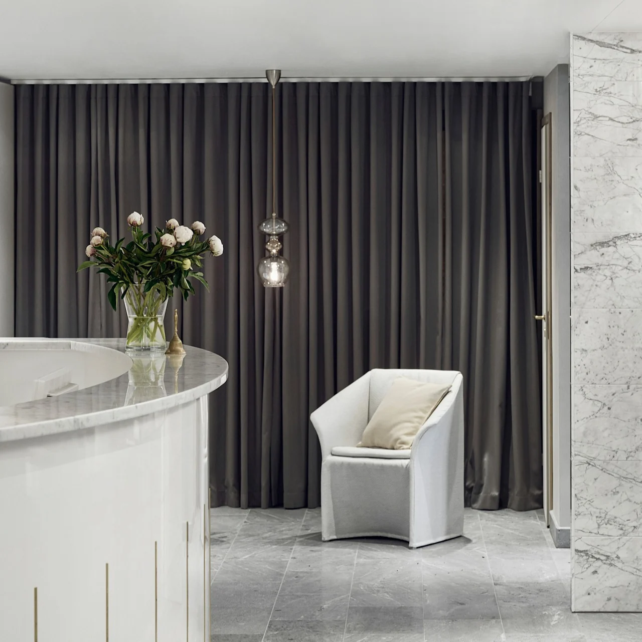 A modern reception area with a curved white desk, hanging glass lights, and a lone chair, against a backdrop of gray curtains and marble accents at Kämp spa in Helsinki.