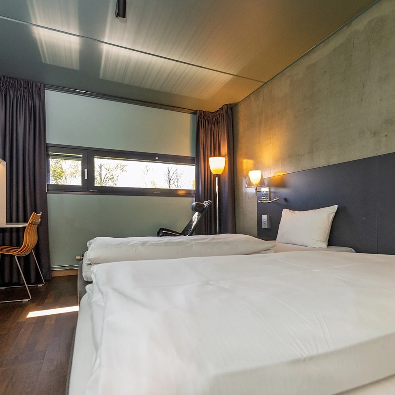 Two neatly made beds rest in a modern hotel room with wooden floors. A desk with a chair sits by a window with curtains, illuminated by wall-mounted lamps.