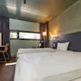 Two neatly made beds rest in a modern hotel room with wooden floors. A desk with a chair sits by a window with curtains, illuminated by wall-mounted lamps.