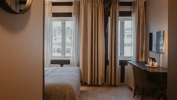 Et hyggeligt hotelværelse med en seng, store vinduer med lyse gardiner, et skrivebord med en stol og et vægmonteret TV.