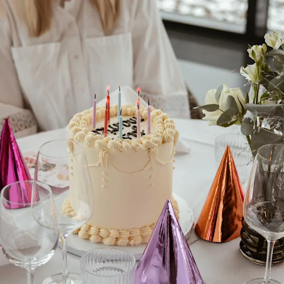 En bursdagskake med tente lys på et festdekket bord, omgitt av festhatter og glass.