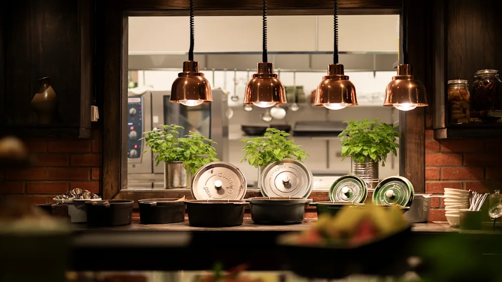 A warm and inviting food service area with a glimpse into a busy kitchen, featuring copper lamps and fresh plants.