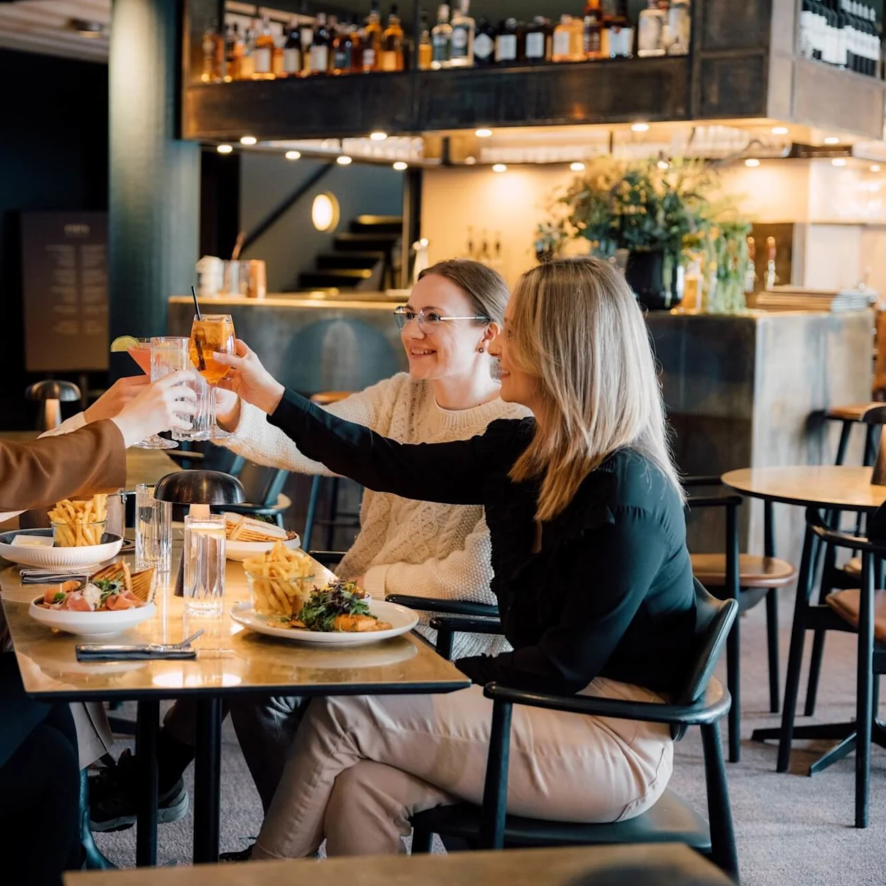 Fire personer sidder ved et restaurantbord, klinker glas i en festlig skål. Tallerkener med mad og glas omgiver dem i et varmt belyst spiseområde.