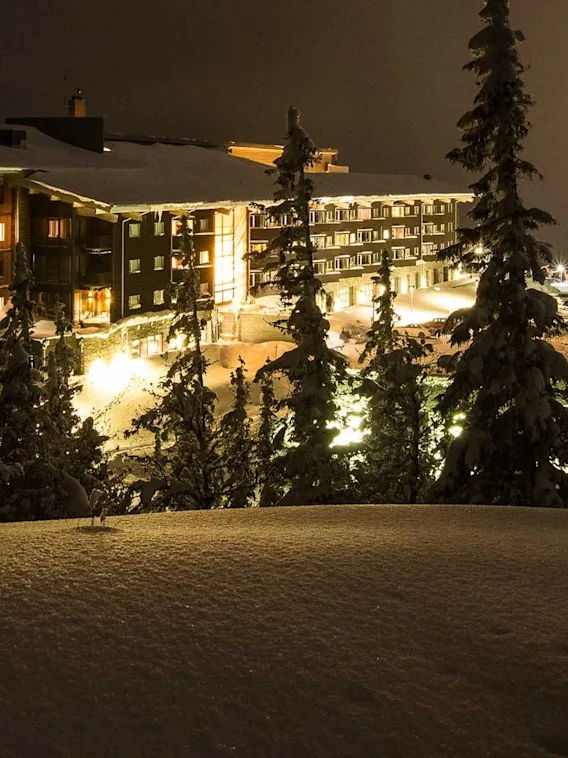 En snötäckt lodge avger varmt ljus på natten bland mörka barrträd, vilket skapar en mysig, fridfull bergsmiljö.