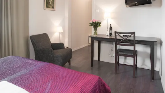 Ett snyggt ordnat hotellrum med en säng, fåtölj, skrivbord och väggmonterad TV i en ren, modern miljö.
