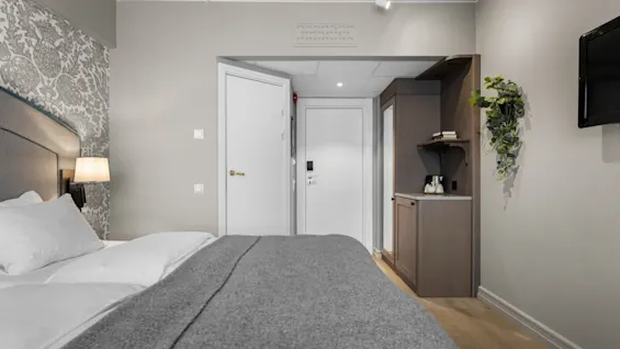 A cozy hotel room with a bed, bedside lamp, and patterned wallpaper. A small cabinet with shelves and a hanging plant adds to the inviting atmosphere.