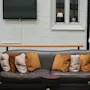 En brun lädersoffa med beige och bruna kuddar står framför en texturerad vit vägg. I närheten finns en skylt som säger "Garderobe & Vinkällare." Växter och en lampa skapar en mysig atmosfär.