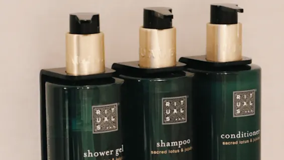 Three dark green bottles labeled "RITUALS": shower gel, shampoo, conditioner, sacred lotus & jujube, mounted on a tiled wall. "THE RITUAL OF JING" noted below with 10.1 FL. OZ / 300 mL.