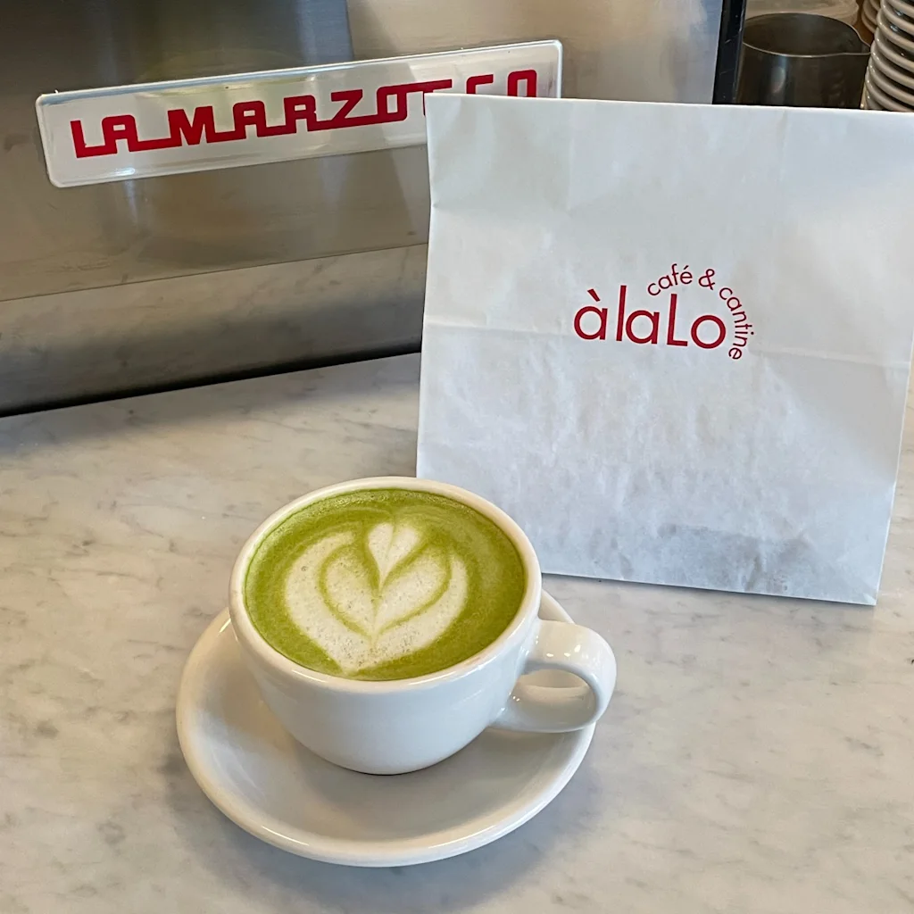 Njut av en livlig grön latte med hjärtformad skumkonst, bredvid en vit påse från cafe & cantine a la Lo, på en kafédisk.