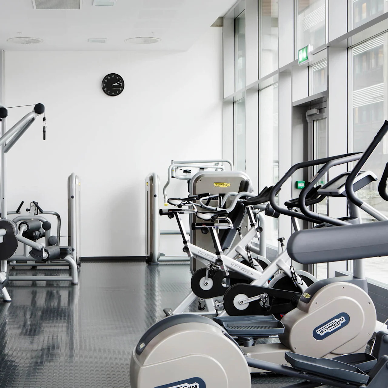 Treningsutstyr er pent ordnet i et moderne, lyst rom med store vinduer og et veggmaleri. Merket Technogym er synlig på flere maskiner, ved siden av en veggklokke.