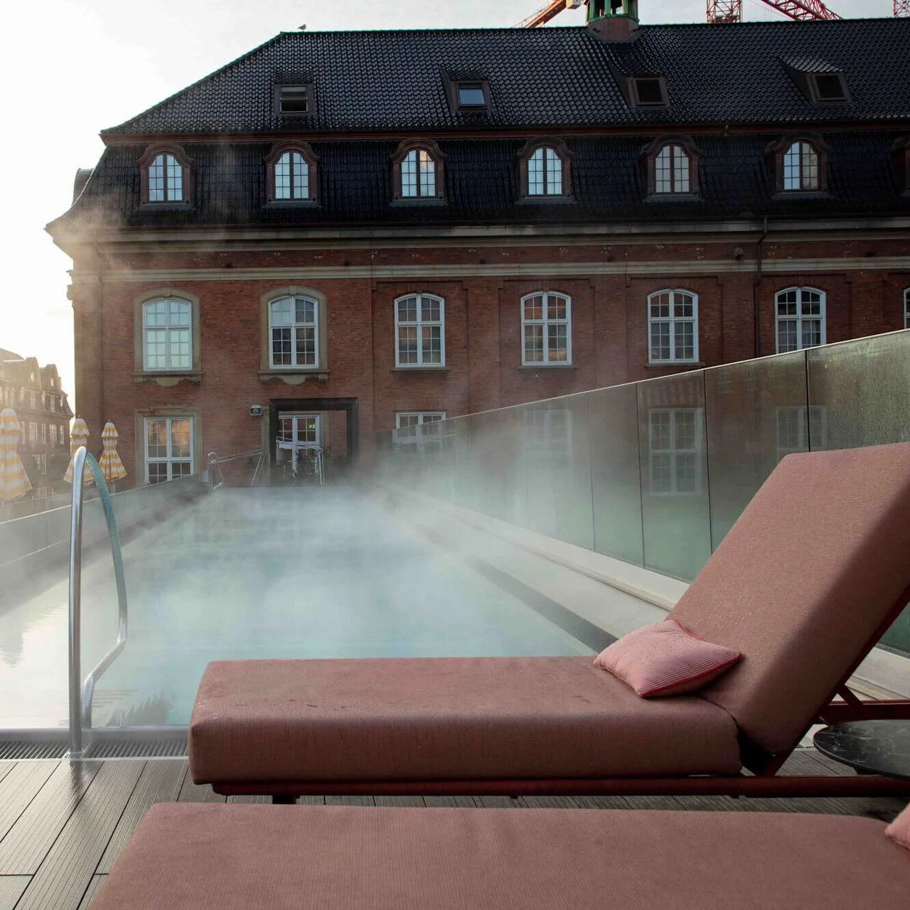A steaming rooftop pool is surrounded by glass railings and loungers, with a historic brick building and striped umbrellas in the background.