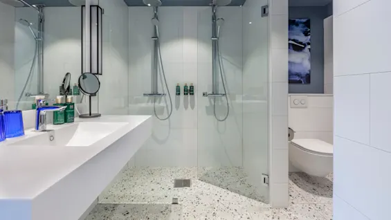 A modern bathroom with a double shower, sink, and toilet. Clean, white-tiled design with transparent shower doors and a terrazzo floor.