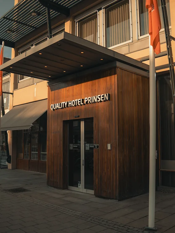 En hotellinngang med skiltet "QUALITY HOTEL PRINSEN" på en trefasade, ligger langs en stille gate med en annen bygning merket "NAPOLI" i nærheten.