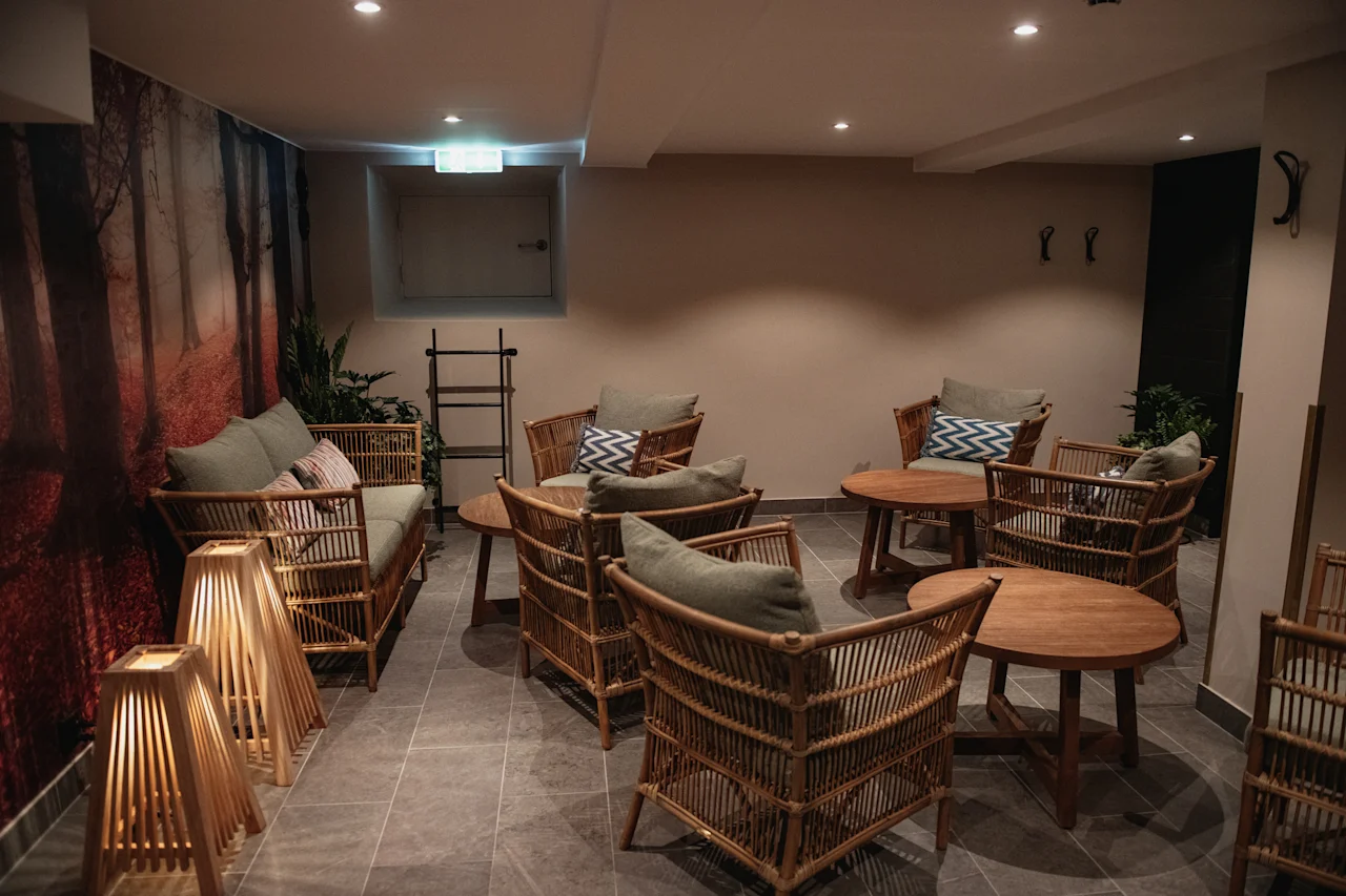 Et koselig salongområde med kurvmøbler, myke puter og trebord, med et naturinspirert veggmaleri og varm belysning.