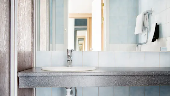 A bathroom sink with a metallic faucet is attached to a speckled countertop. A mirror reflects an open door and towel, with light blue tiles in the background.