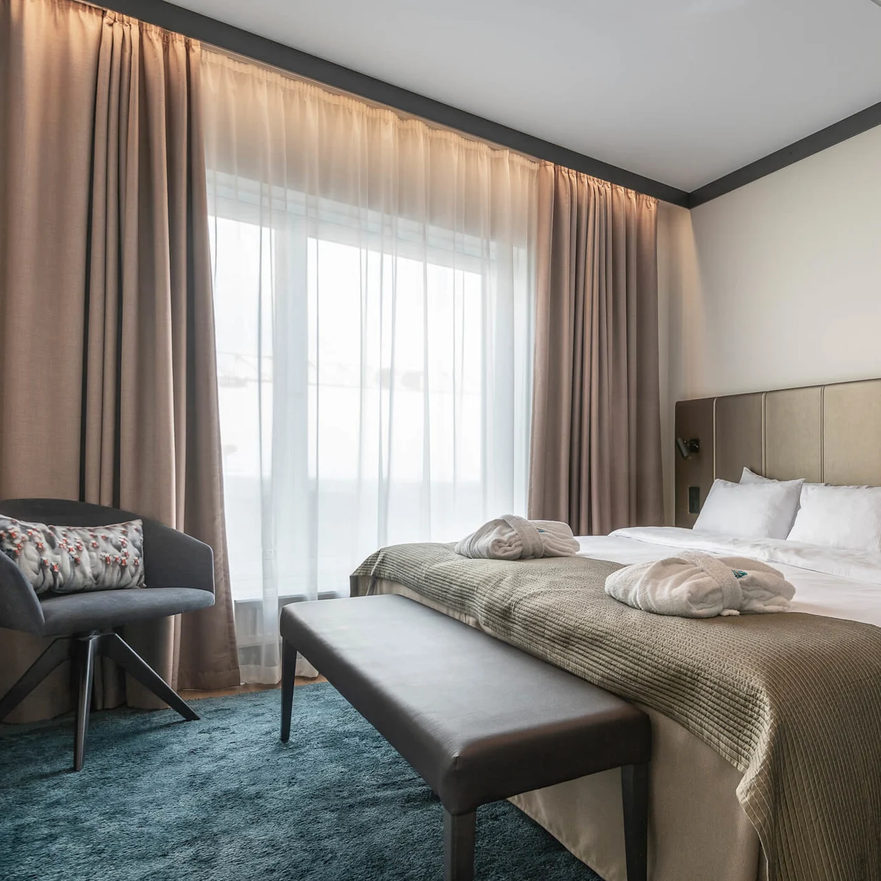 A neatly made bed with pillows and robes sits beside a window with beige curtains, surrounded by a chair, bench, and soft blue carpet in a cozy room.