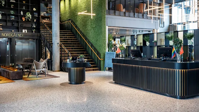 Reception desk adorned with flags stands in a modern lobby; lush green wall and stairs in the background. Text includes "NORDGÅENDE."