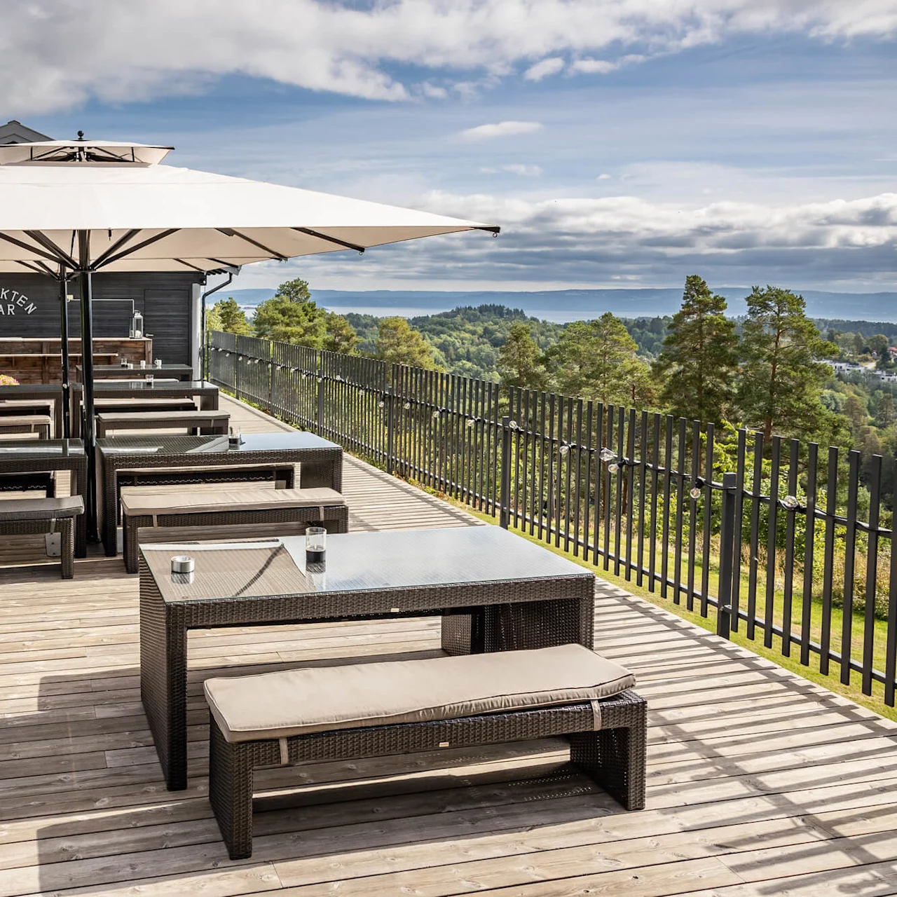 Uteterrasse med rottingmøbler under store parasoller; "UTSIKTEN BAR" på trebart. Utsikt over frodig grønt landskap og fjerne åser under en delvis skyet himmel.